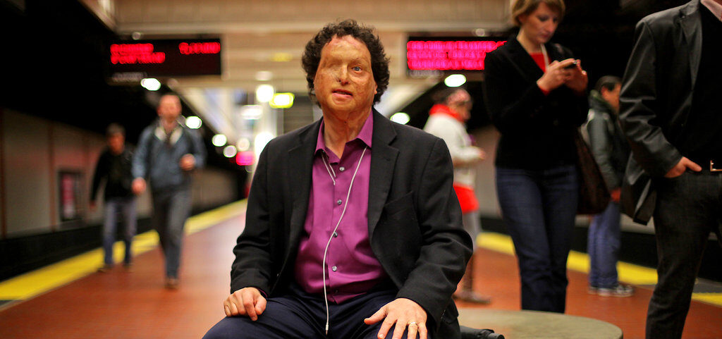 Josh Miele sits in the middle of a subway terminal. He is in sharp focus while the background and others around him are blurred.